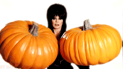 Elvira, Mistress of the Dark, balancing one enormous pumpkin on each hand.